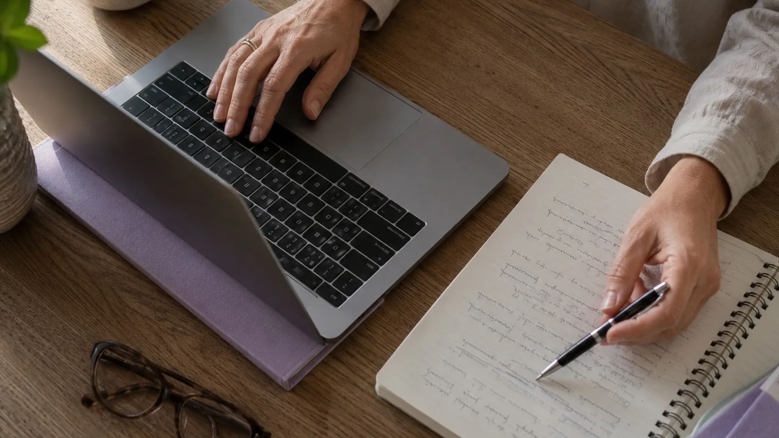 A person's hands at a laptop on a timber desk with a notepad and pen beside it